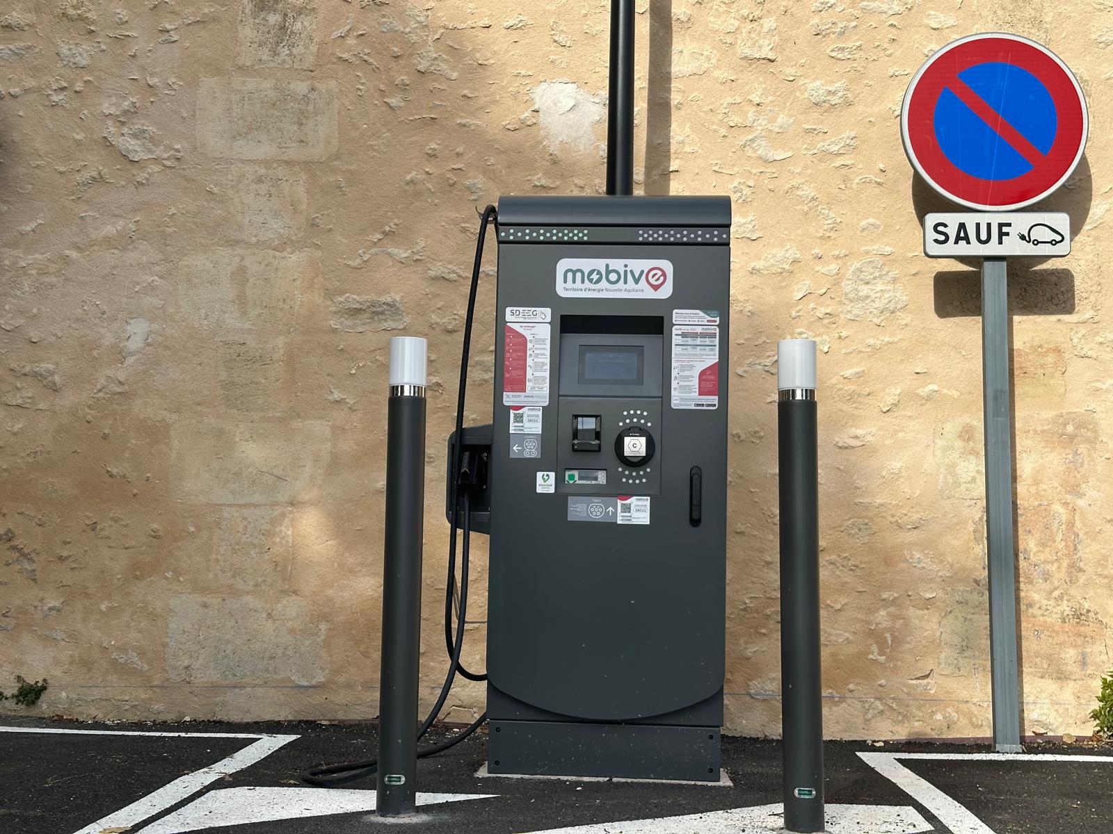 IMG-20251031-WA0010 Borne de recharge pour véhicules électriques Mobive installée contre un mur en pierre, avec un panneau indiquant “stationnement interdit sauf véhicules électriques”.